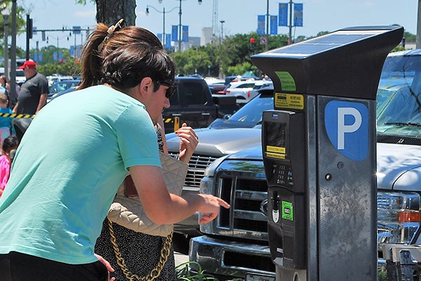 A man and woman use a parking meter on a sunny day. Cars are parked in the background.