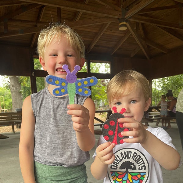 Two children stand under a wooden shelter, smiling and holding felt crafts of a butterfly and a ladybug.